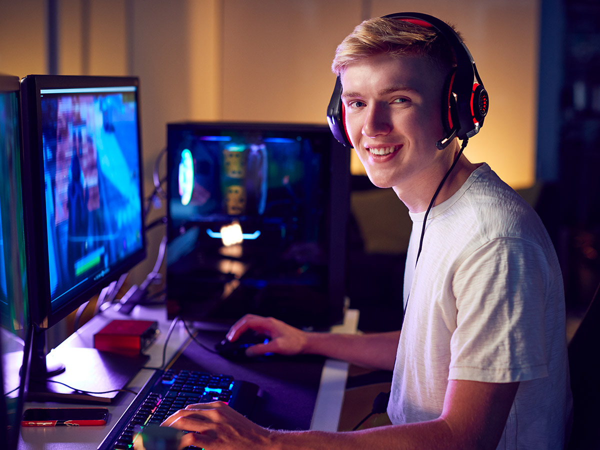 Image of a young man wearing a headset, sitting in front of a gaming computer with dual screens